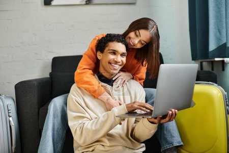 alegre pareja multiétnica de compras en línea, la celebración de una tarjeta de crédito, con un ordenador portátil cerca de equipaje