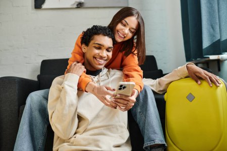 Photo for Content woman embraces her black boyfriend on a cozy couch and holding smartphone near him - Royalty Free Image