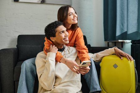 Photo for Happy woman embraces her black boyfriend on a cozy couch and holding smartphone near him, travel - Royalty Free Image