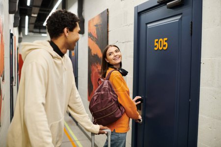 Cheerful woman with backpack unlocking her hostel room door, looking at african american boyfriend