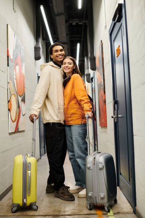 Feliz pareja diversa en un corredor de albergue con maletas, disfrutando de una experiencia de viaje alegre