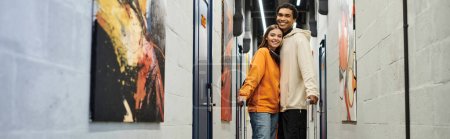 Relaxed multicultural couple with luggage smiling and standing together in a hostel hallway, banner