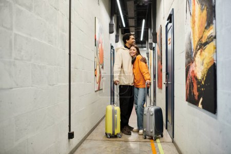 Relajada pareja multicultural con equipaje abrazando y de pie juntos en un pasillo albergue