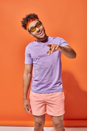 happy young african american man in eyeglasses pointing with finger on orange background, gesture