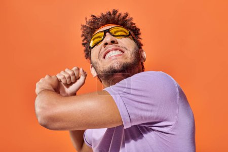 verspielter junger afrikanisch-amerikanischer Mann gestikuliert und lächelt auf orangefarbenem Hintergrund, dynamische Bewegung