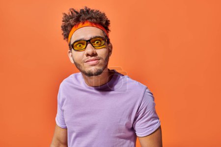 Téléchargez les photos : Drôle de visage, jeune homme afro-américain en lunettes de soleil en regardant la caméra sur fond orange - en image libre de droit