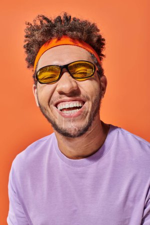 Téléchargez les photos : Heureux homme afro-américain en lunettes souriant avec les yeux fermés sur fond orange, optimiste - en image libre de droit