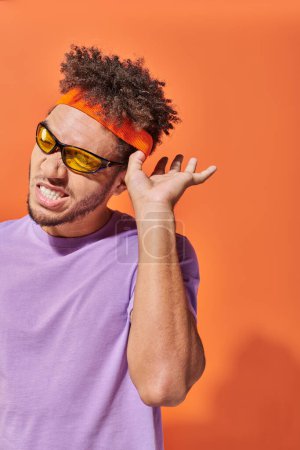 neugieriger afrikanisch-amerikanischer Mann mit Sonnenbrille gestikuliert beim Zuhören auf orangefarbenem Hintergrund