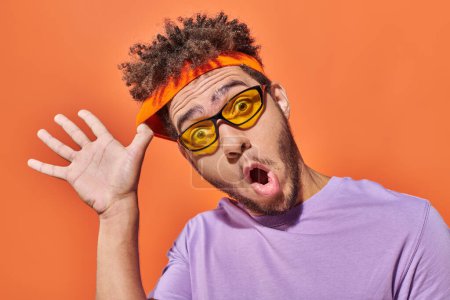 funny african american man in sunglasses adjusting headband on orange background, surprised face