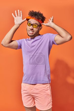 joven afroamericano hombre en gafas de sol ajustando diadema sobre fondo naranja, cara expresiva