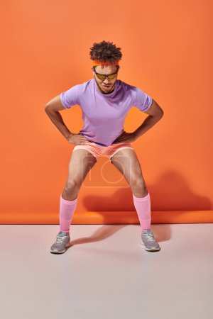 sportlicher afrikanisch-amerikanischer Mann in Sonnenbrille und Turnbekleidung beim Training auf orangefarbenem Hintergrund