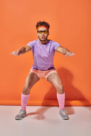 sportlicher afrikanisch-amerikanischer Mann in Sonnenbrille und Turnbekleidung hockt auf orangefarbenem Hintergrund