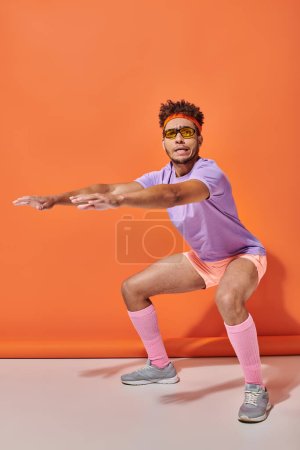 junger afrikanisch-amerikanischer Sportler in Sonnenbrille und Turnbekleidung hockt auf orangefarbenem Hintergrund