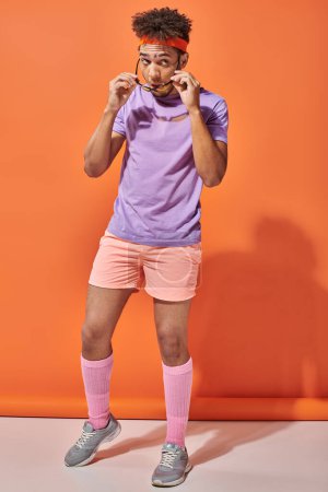 young african american sportsman in gym attire wearing sunglasses on orange background, fitness