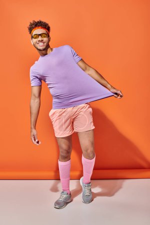 cheerful african american sportsman in gym attire showing his purple t-shirt on orange background