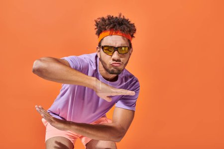 Selbstbewusster afrikanisch-amerikanischer Sportler in Turnbekleidung und Sonnenbrille gestikuliert auf orangefarbenem Hintergrund