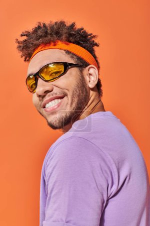 cheerful african american man in eyeglasses and headband smiling on orange background, optimistic mug #692584724