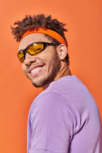 cheerful african american man in eyeglasses and headband smiling on orange background, optimistic hoodie #692584724