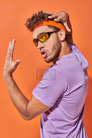 expresivo hombre afroamericano en gafas y diadema mirando a la cámara sobre fondo naranja