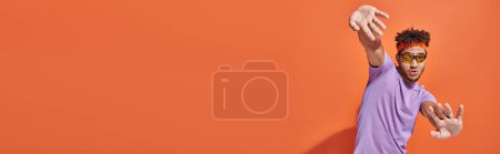 funny african american man in eyeglasses and headband looking at camera on orange background, banner