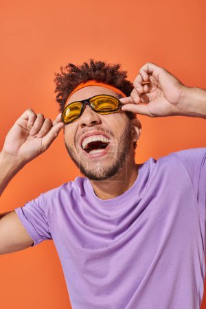 Téléchargez les photos : Heureux afro-américain en bandeau souriant et portant des lunettes de soleil sur fond orange - en image libre de droit