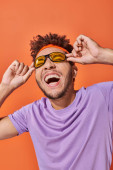 happy african american man in headband smiling and wearing sunglasses on orange background Poster #692585108