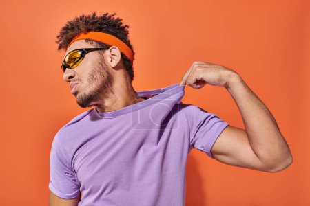 confident african american man in headband adjusting purple t-shirt on orange background mug #692585118