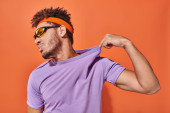 confident african american man in headband adjusting purple t-shirt on orange background Poster #692585118