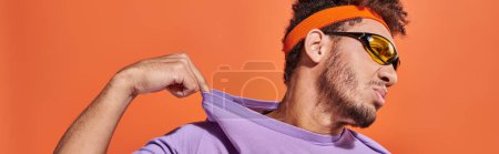 banner of confident african american man in headband adjusting purple t-shirt on orange background Poster 692585124