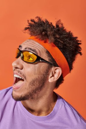 excited african american man in eyeglasses and headband on orange background, open mouth