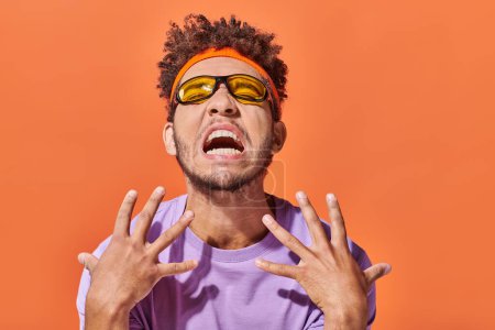 Téléchargez les photos : Homme afro-américain émotionnel en bandeau et lunettes de soleil geste sur fond orange - en image libre de droit