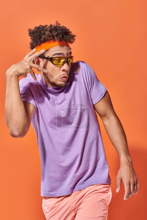 Téléchargez les photos : Surpris homme afro-américain dans les lunettes et bandeau regardant loin sur fond orange, geste - en image libre de droit