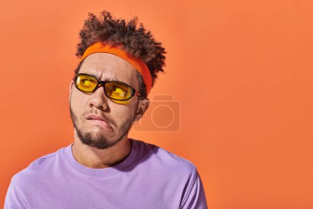 Téléchargez les photos : Cher homme afro-américain en lunettes de soleil et bandeau regardant loin sur fond orange, grimace - en image libre de droit