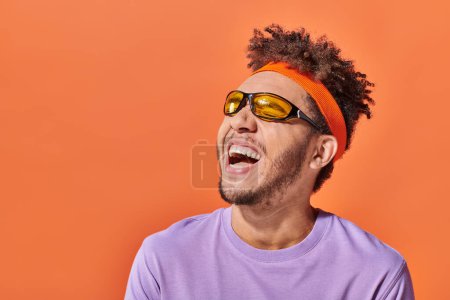Téléchargez les photos : Afro-américain en lunettes de soleil et bandeau riant et regardant loin sur fond orange - en image libre de droit
