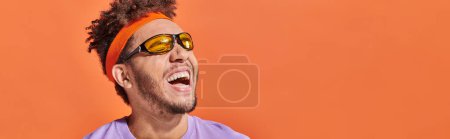 Photo for African american man in sunglasses and headband laughing and looking away on orange backdrop, banner - Royalty Free Image