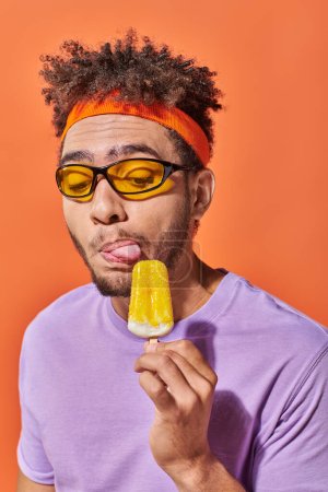 Photo for African american man in sunglasses and headband licking fruity ice cream on orange backdrop - Royalty Free Image