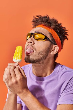 Photo for African american man in sunglasses and headband licking fruity ice cream on orange backdrop, tongue - Royalty Free Image