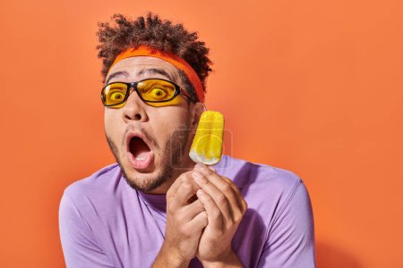 shocked african american man in sunglasses and headband holding frozen ice cream on orange