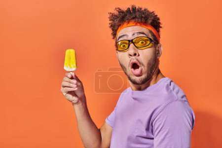 shocked african american man in sunglasses and headband holding frozen ice cream on orange backdrop