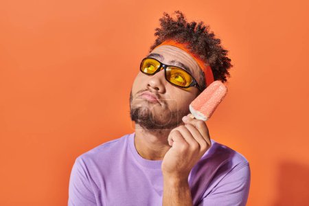 hombre afroamericano pensativo en gafas de sol celebración de helado con aspersiones sobre fondo naranja