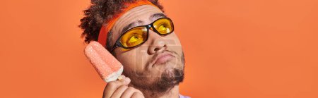 pensive african american man in sunglasses holding ice cream with sprinkles on orange, banner