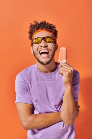 happy african american man in sunglasses holding ice cream with sprinkles on orange backdrop