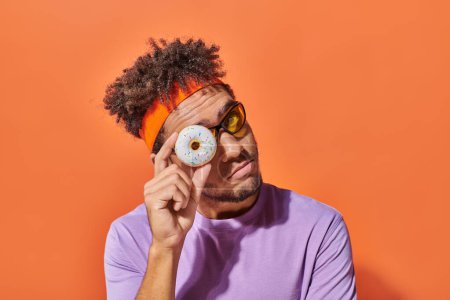 joven africano americano hombre en gafas de sol mirando cámara a través de donut agujero en naranja fondo