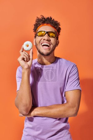 happy young african american man in sunglasses holding small tasty donut on orange background