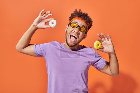 glücklicher junger afrikanisch-amerikanischer Mann mit Sonnenbrille und kleinen leckeren Donuts auf orangefarbenem Hintergrund