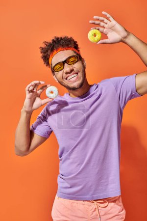 happy young african american man in sunglasses holding bite-sized donuts on orange background