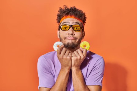 lustige junge afrikanisch-amerikanische Mann mit Sonnenbrille hält mundgerechte Donuts auf orangefarbenem Hintergrund