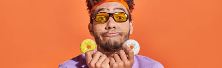 funny african american man in sunglasses holding bite-sized donuts on orange background, banner