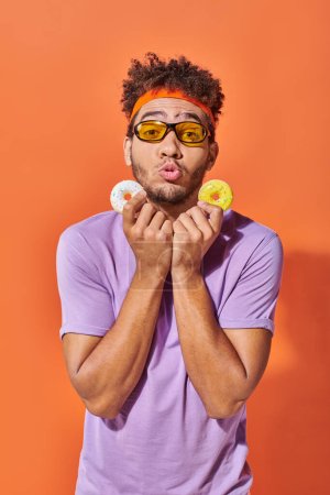 young african american man in sunglasses holding bite-sized donuts on orange background, grimace