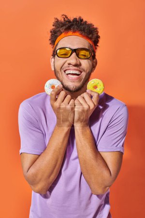 joyful young african american man in sunglasses holding bite-sized donuts on orange background